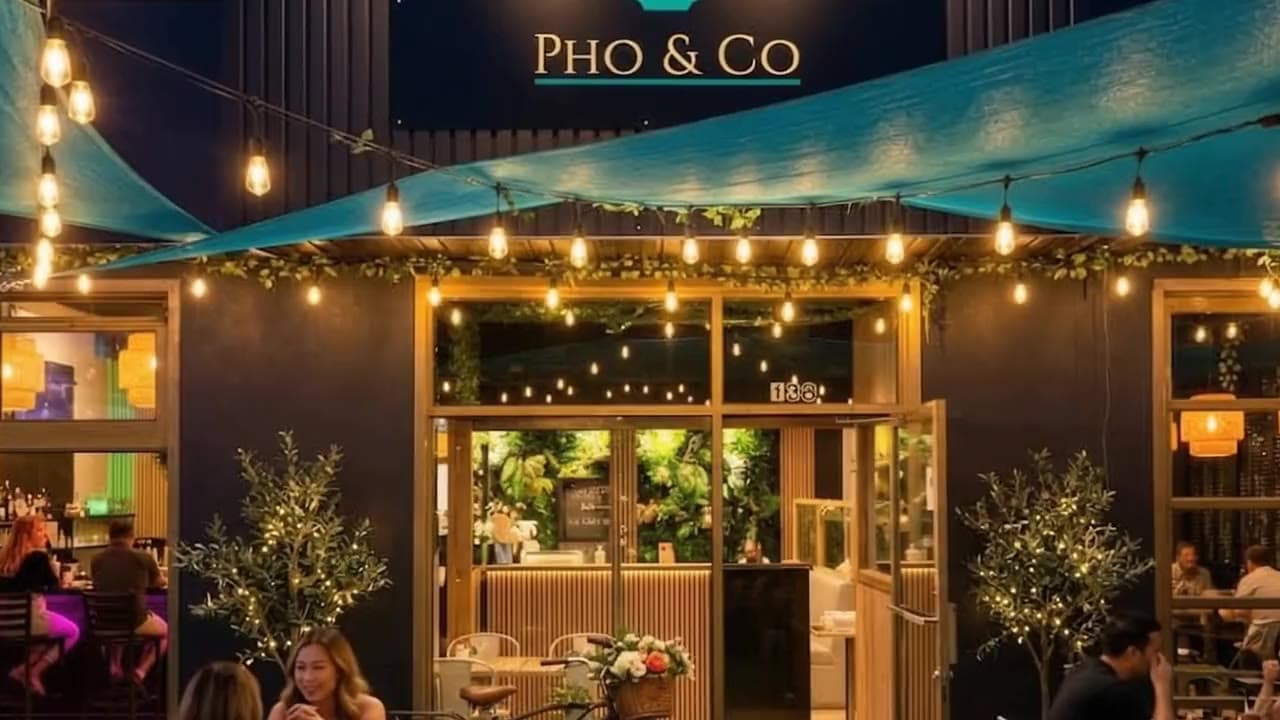La façade de Phở & Co. le soir : l’enseigne, les guirlandes, un couple qui entre sur la terrasse